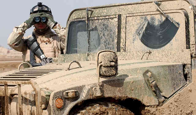 Image of a soldier peering through binoculars while standing next to a mud-caked military vehicle